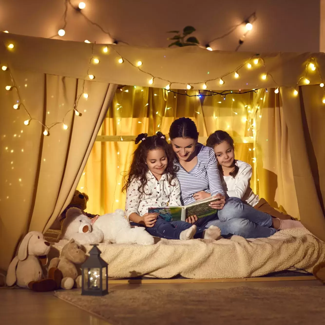 A mother reading a book to her two daughters in front of an indoor fort with sheets and twinkly lights.